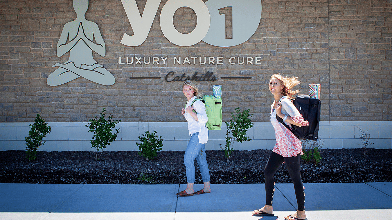 Health & Wellness Camp – Rebalance From Within at YO1 Longevity & Health Resorts - two guests carrying backpacks walking past the YO1 Luxury Nature Cure sign at the resort