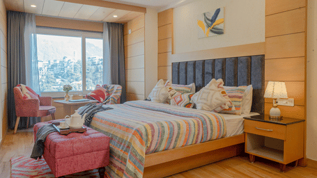 A bedroom with a king size bed, patterned bedding, bedside tables, a bench, 2 chairs, a lamps, wooden flooring, and a window at Perfectstayz Premium At Mall Road (Hotel Super), Mussoorie.