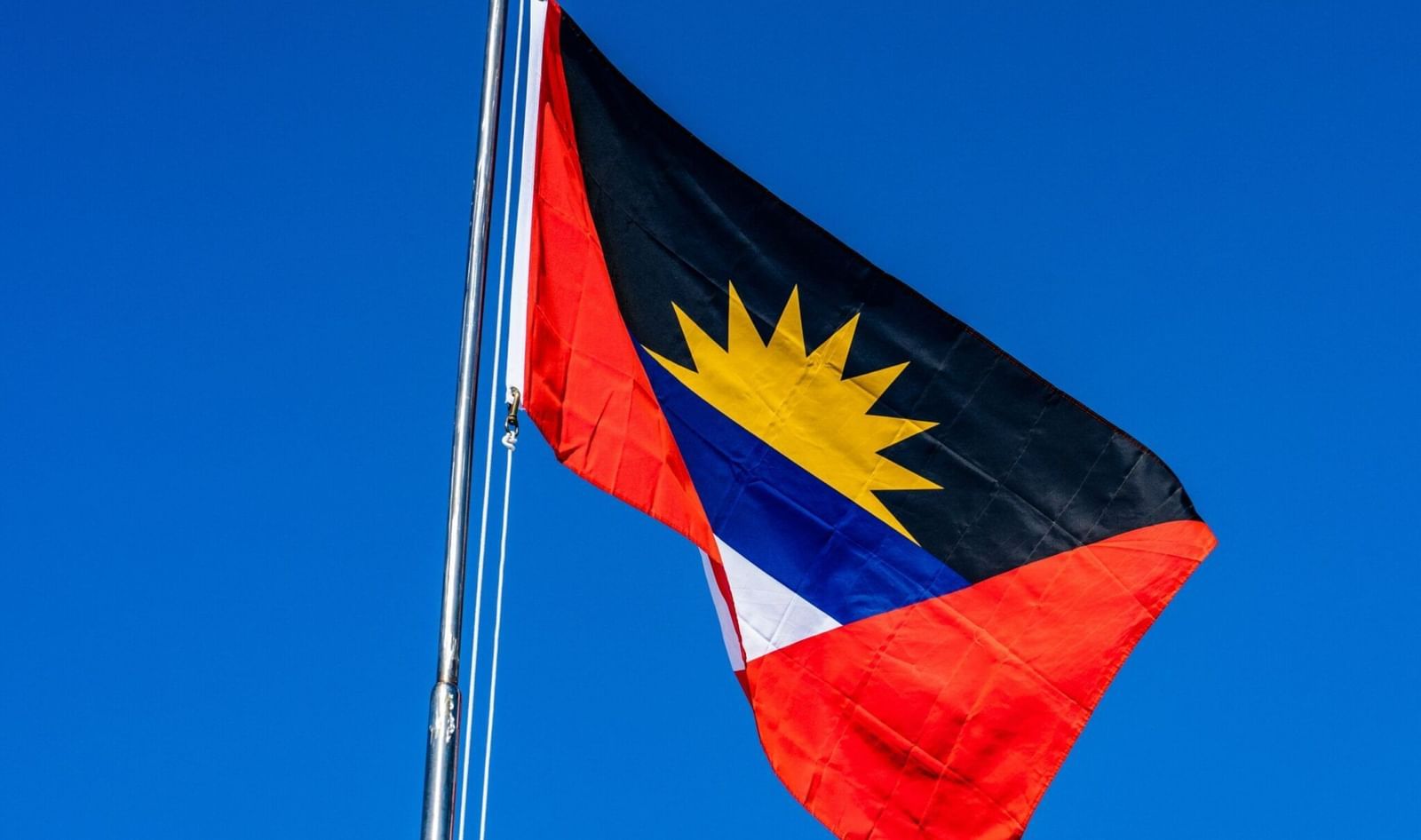 The flag of Antigua and Barbuda, featuring a black triangle with a rising yellow sun over a blue band, flanked by red triangles on either side, flying against a clear blue sky.