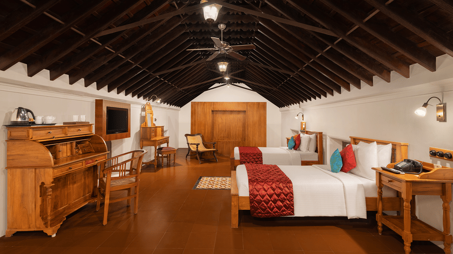 A spacious bedroom at Chidambara Vilas - A Luxury Heritage Resort, with a twin beds, tiled flooring, wooden beams on the ceiling, and a wall mounted television.
