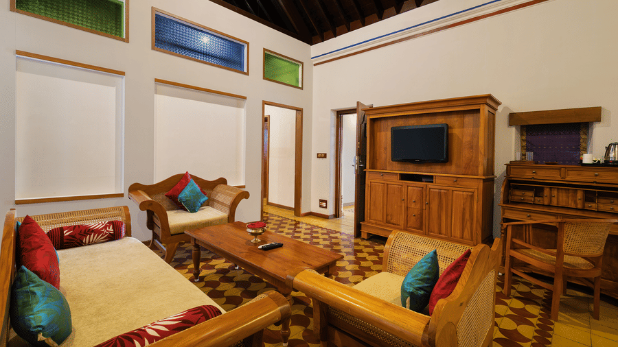 Living area in a hotel room at Chidambara Vilas - A Luxury Heritage Resort, featuring wooden furniture, a dining table with chairs, a sofa, and a television cabinet.