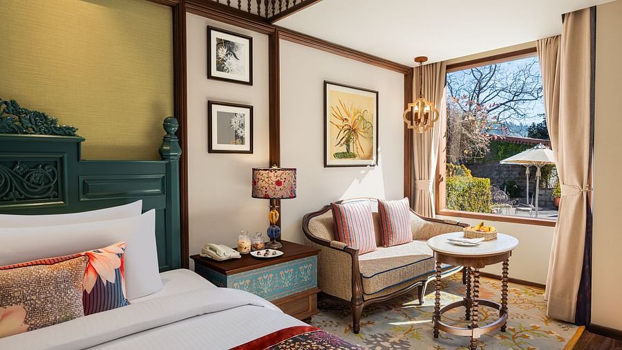 An image of a premium garden room in the naini retreat, nainital, with a king-size bed, wooden furniture, and a bay window overlooking a garden.  and a small sitting area, with a coffee table and chair along window side.