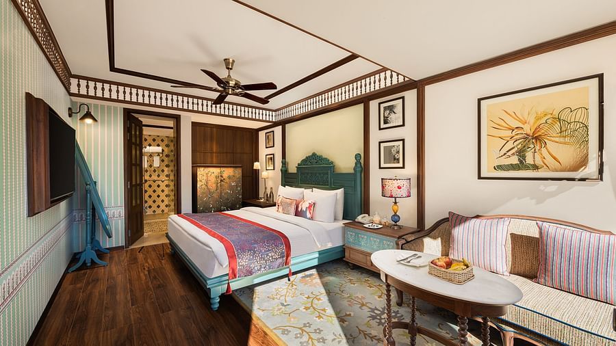An image of a premium garden room in the naini retreat, nainital, with a king-size bed, wooden furniture, and a bay window overlooking a garden. The room features a ceiling fan, a television, and a small sitting area. 
