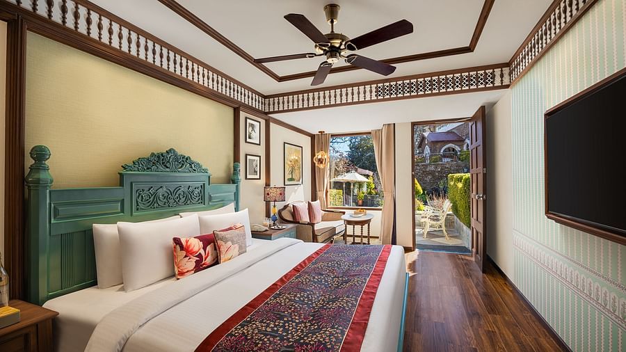 An image of a premium garden room in the naini retreat, nainital, with a king-size bed, wooden furniture, and a bay window overlooking a garden. The room features a ceiling fan, a television, and a small sitting area. 