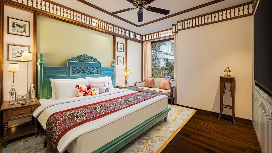 An image of a  cozy room in the naini retreat, nainital, with a king-size bed, wooden furniture, and a bay window overlooking a garden. The room features a ceiling fan, a television, and a small sitting area. 