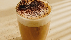  tall glass of layered coffee with a thick layer of foam and a dusting on top, accompanied by a few coffee beans on a light surface at  Gemini Continental