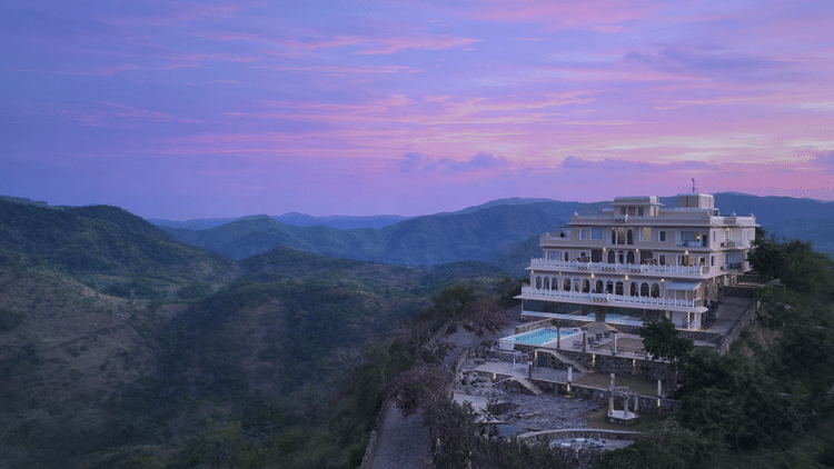 Facade of Fateh Safari Suites, Kumbhalgarh featuring multiple storeys, swimming pool, lawn, sitting area, and more amidst the green hills.