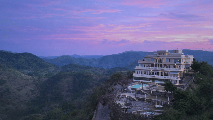 Facade of Fateh Safari Suites, Kumbhalgarh featuring multiple storeys, swimming pool, lawn, sitting area, and more amidst the green hills.