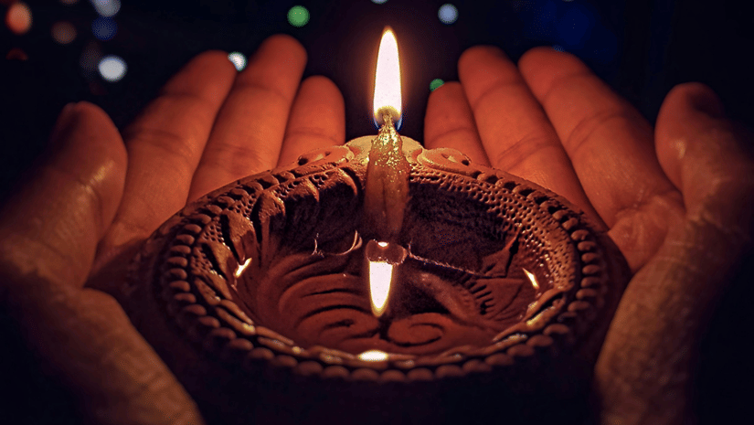 A person's hands cupping a traditional clay lamp with a single, bright flame in the center. Importance of Diwali include lighting a diya to signify light over darkness.
