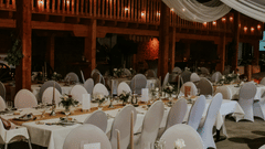 A view of a decorated indoor event space, possibly a wedding reception, with round tables set with white tablecloths, chairs, and elegant lighting.