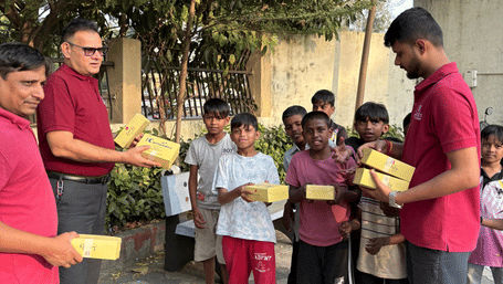 Group of men in red shirts distributing food boxes to smiling children outdoors near trees and a wall, during a community outreach activity.