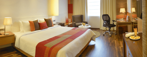 A room at Deventue Sarovar Portico - Kapashera, New Delhi featuring a double size bed, a work station, a sofa chair, bedside table on either side, a cabinet table, and a window.