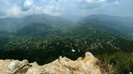 An overview of Chauli Ki Jali, Mukteswar, with a rocky surface in the foreground and a valley in the distance with blue sky in the background.