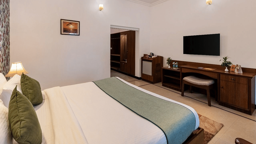 A bedroom at Amantra Shilpi Resort, Udaipur showing a king size bed with green cushions, a wall-mounted television, desk, and an open doorway.