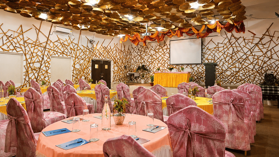 Banquet hall with round tables dressed in yellow and peach linen, pink patterned chair covers, a decorative bamboo ceiling and a stage backdrop.