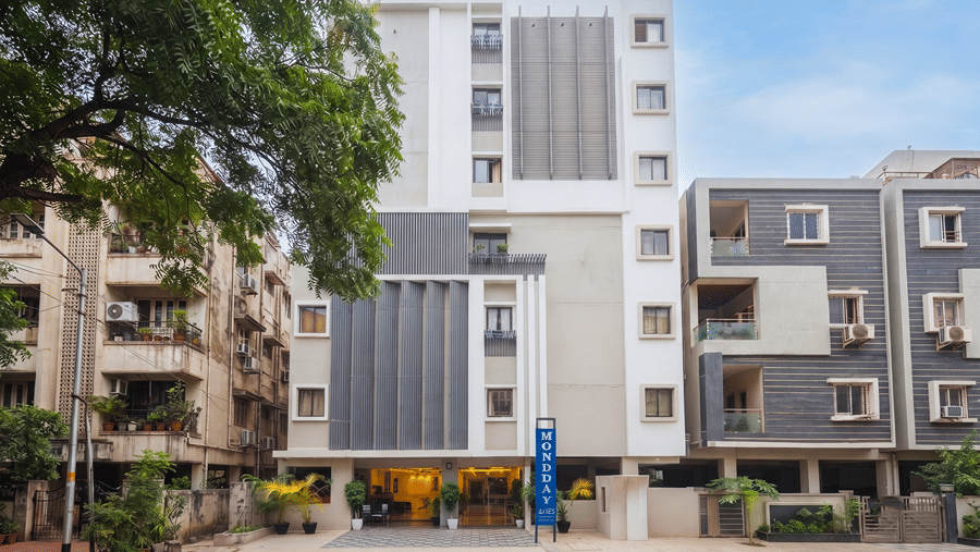 Front facade of Monday Hotels & Suites Madhapur, Hyderabad in daylight, with a blue vertical sign, lit entrance, potted palms, trees and neighbouring buildings.