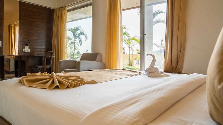 A hotel bedroom with a neatly made bed, featuring two towels folded into swan shapes, and a window offering a view of outdoor greenery - Pride Premier Dehradun