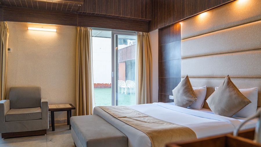 A bright hotel bedroom featuring a large bed with a padded headboard, an armchair in the corner, and warm lighting - Pride Premier Dehradun