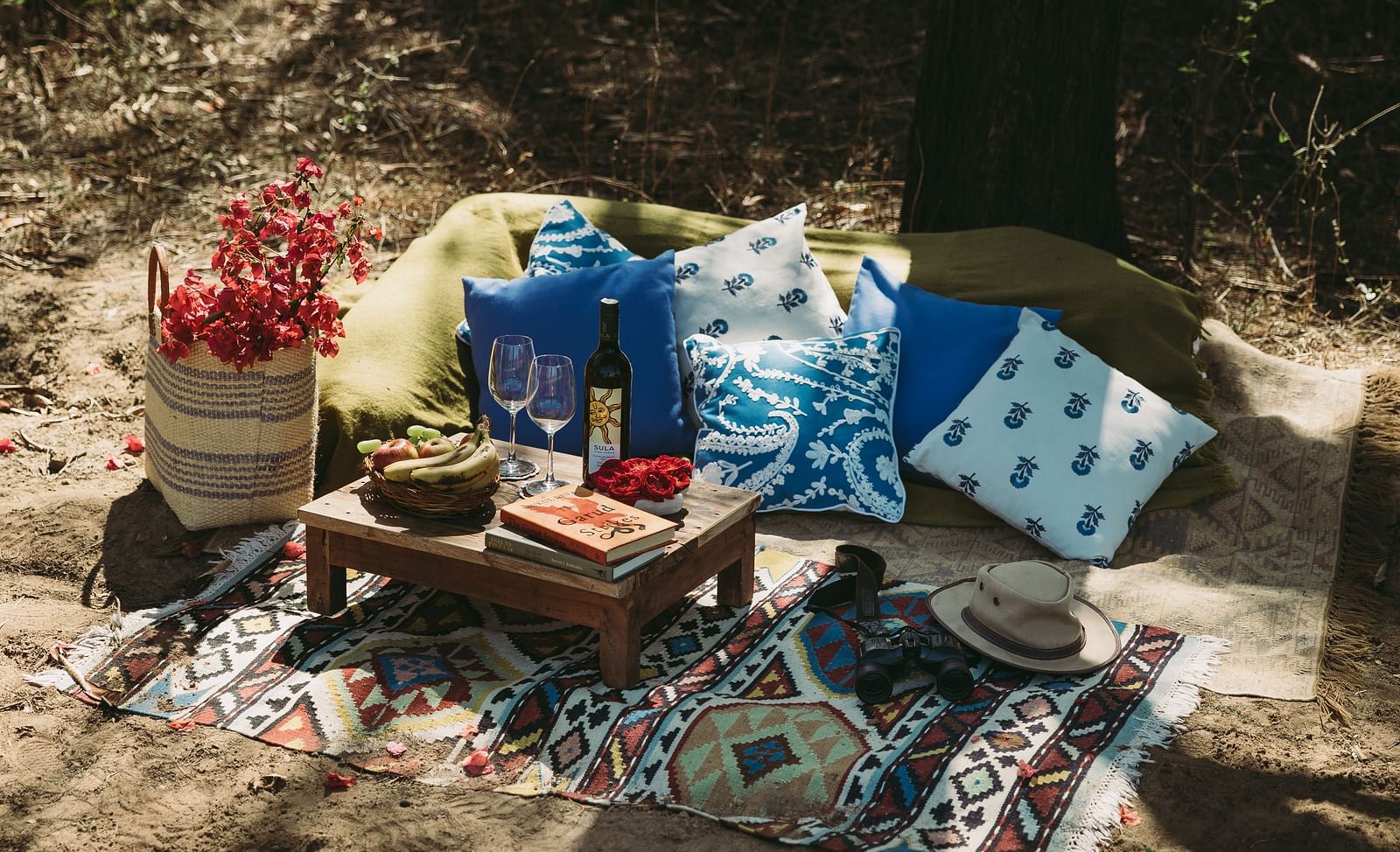 A picnic setup on a sandy surface with a small wooden table, pillows, a blanket, a basket with flowers, and a hat- Shahpura Bagh. 
