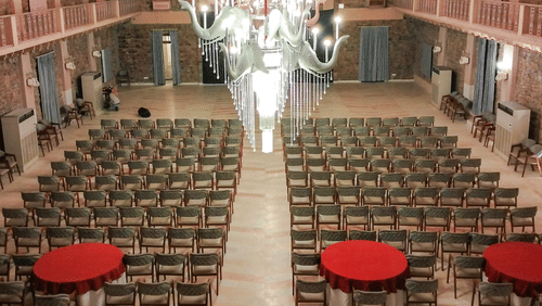 A conference hall at Tijara Fort-Palace - 19th Century, Alwar featuring seating arrangements with chairs and round tables under a well-lit chandelier hanging from a patterned ceiling
