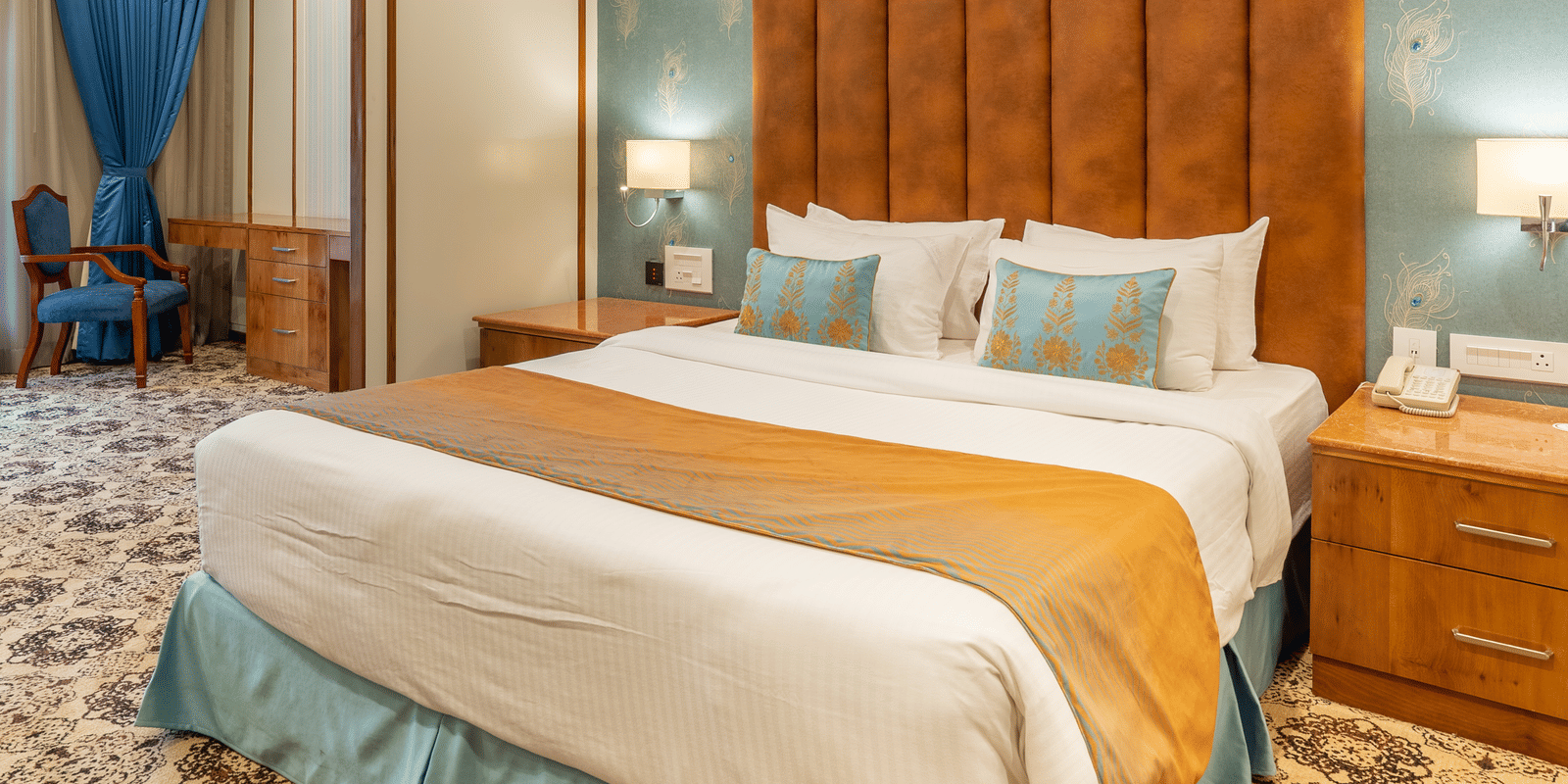 King size bed with blue and orange accents, a large, tufted orange headboard, and ornate patterned carpet at Hotel Hukam's Lalit Mahal.