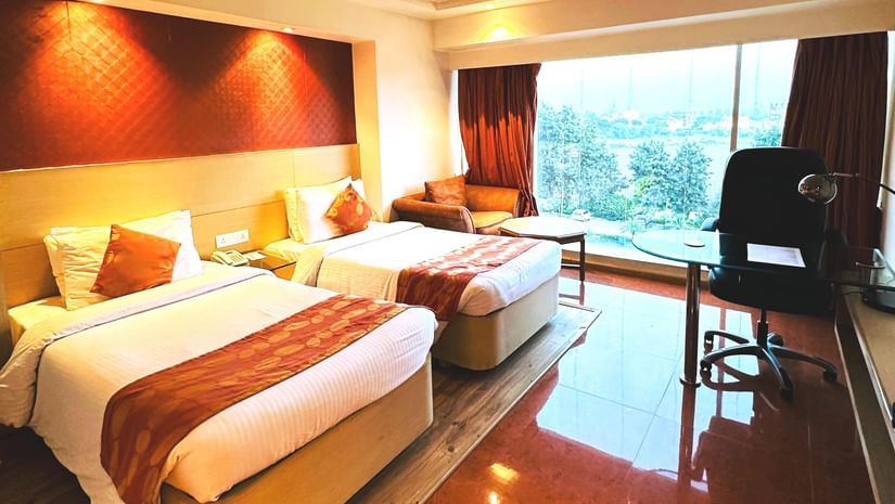  A hotel room at Amer Greens featuring two beds with orange bed runners, a dark headboard, and a large window overlooking the outdoors.