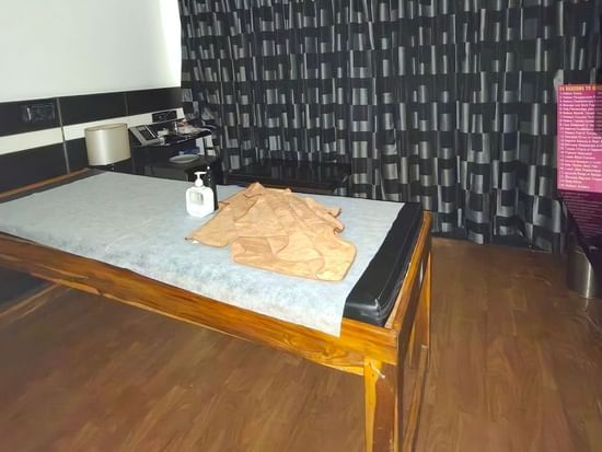 A clean, simple massage treatment room with a wooden massage table, wooden floor, and a dark patterned wall.