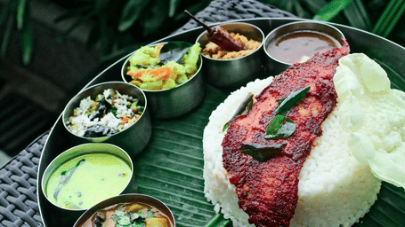 South Indian fish thali on a banana leaf with red masala fish, steamed rice, coconut chutney, sambar, rasam and pickle in small bowls.