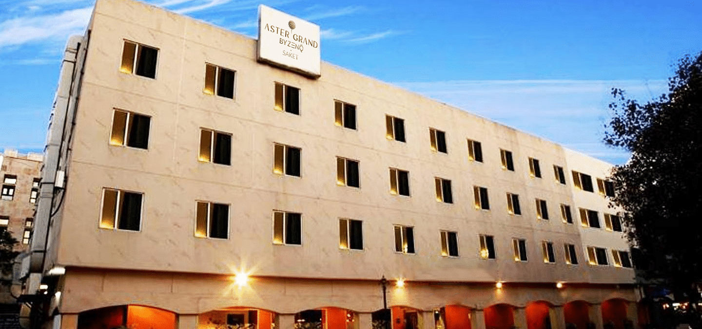  Front view of ASTER GRAND BY ZENQ SAKET, a modern beige hotel with uniform windows and a bright blue sky above.