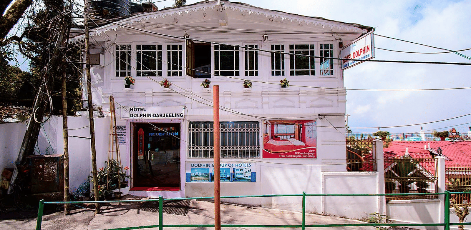 A 2-storey white guesthouse with a green railing and a light blue sky in the background at Hotel Dolphin Darjeeling.