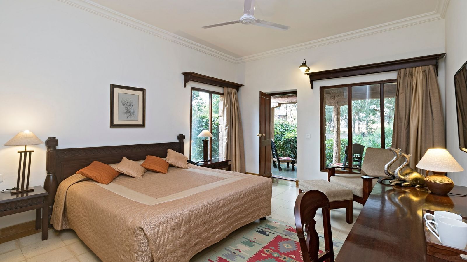 A bedroom with a double bed, side tables with lamps, desk with cups and decor, wooden chairs, patterned rug, ceiling fan, and doors leading outside - MANVÂR Shergarh The Desert Resort