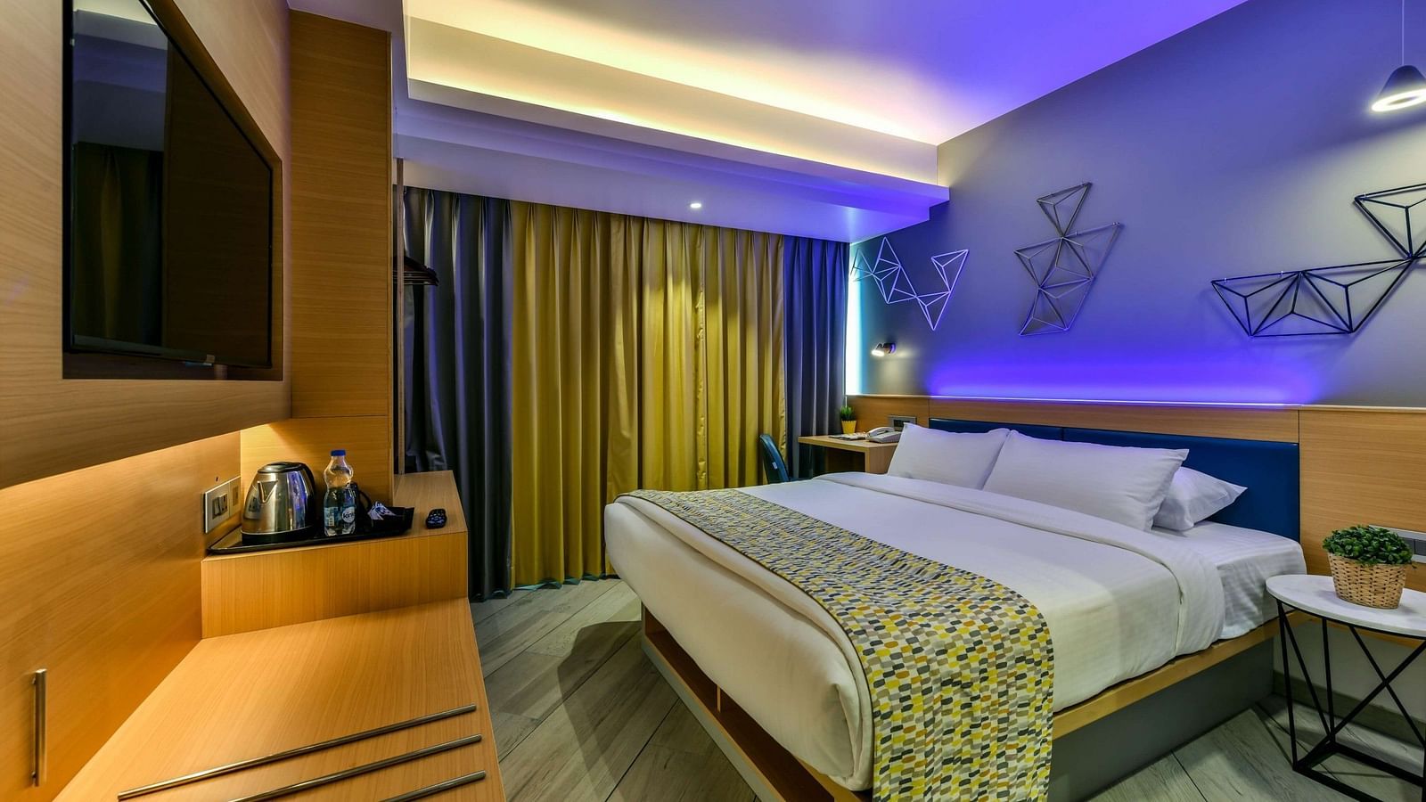 A spacious room with a large bed, a wall-mounted television, a desk positioned below it, and windows with curtains drawn, illuminated by cool lighting at Indie Stays, BKC Kalina.