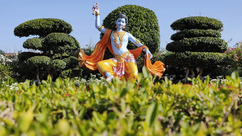 Statue of a Hindu deity (Krishna) seated amongst manicured green topiary and shrubs.