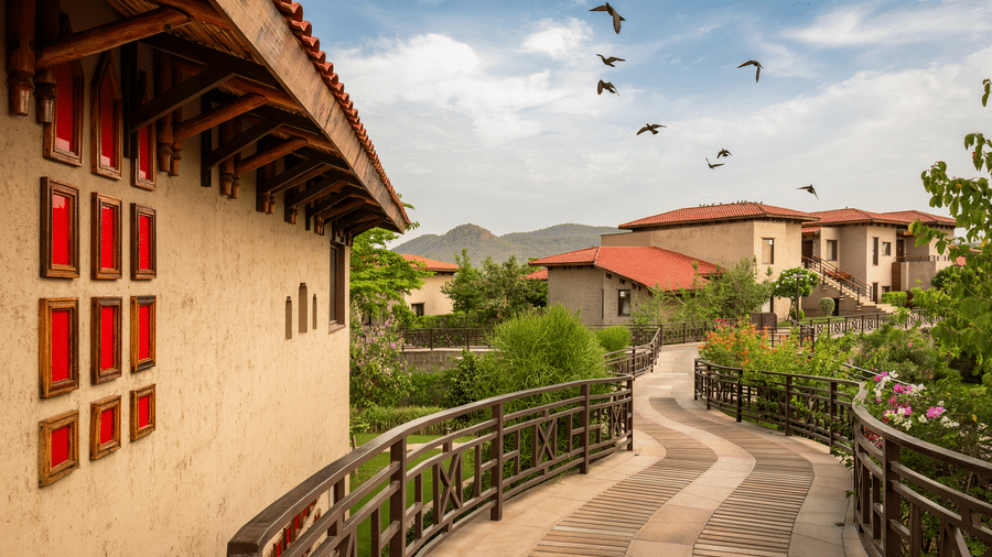 Curving elevated stone walkway framed by rustic wooden railings and red-accented wall niches at AamaGhati Wildlife Resort, set against resort cottages and a flock of birds in the sky.