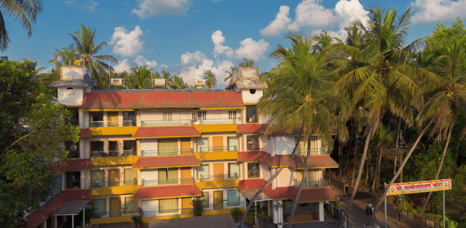 A colourful, low-rise hotel complex with many windows, surrounded by lush green palm trees under a bright sky - Perfectstayz Premium Goa Continental