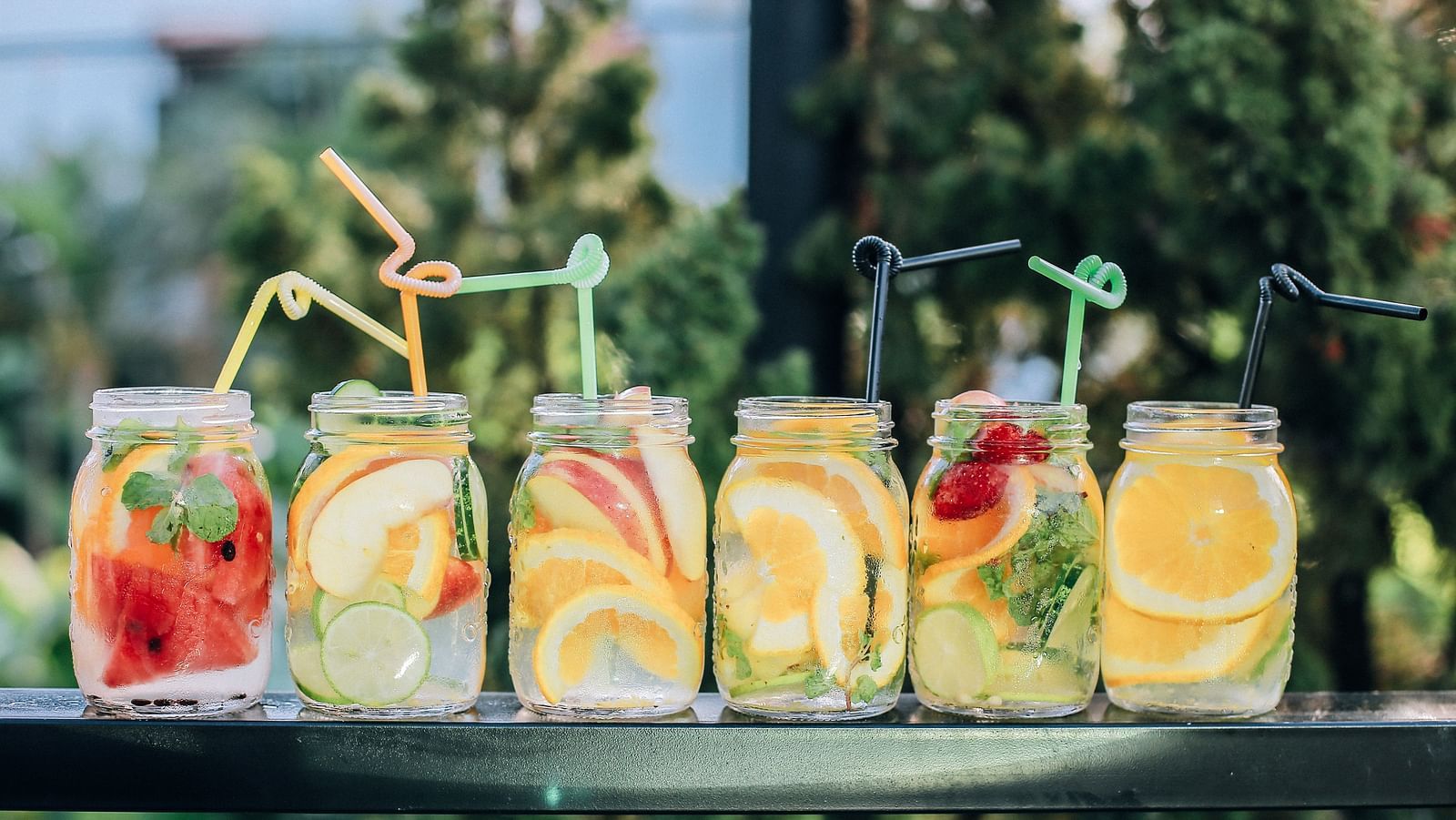 Many drinks kept in a row with fruits inside them and trees in the background being blurred