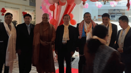 Group of men, including a Buddhist monk, standing at a formal event entrance decorated with red and pink balloons and drapery.
