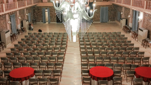 A conference hall at Tijara Fort-Palace - 19th Century, Alwar featuring seating arrangements with chairs and round tables under a well-lit chandelier hanging from a patterned ceiling