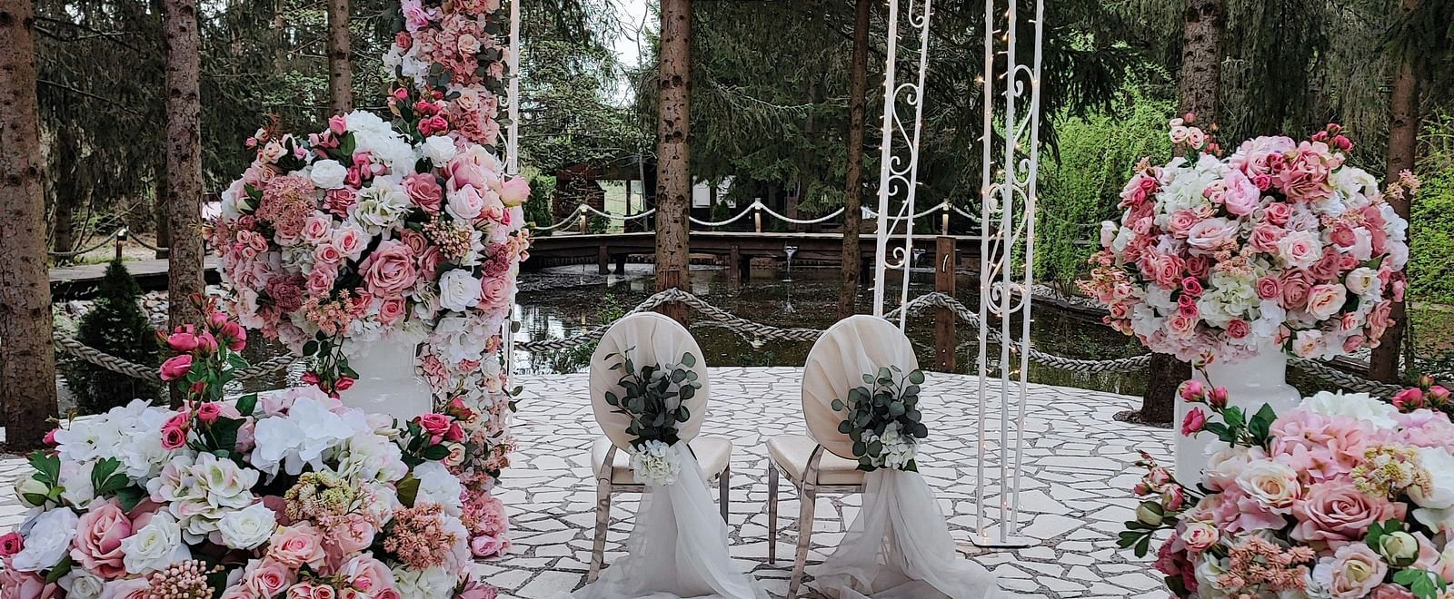 Two white coloured chairs kept under a gazebo decorated with flowers surrounded by dense trees.