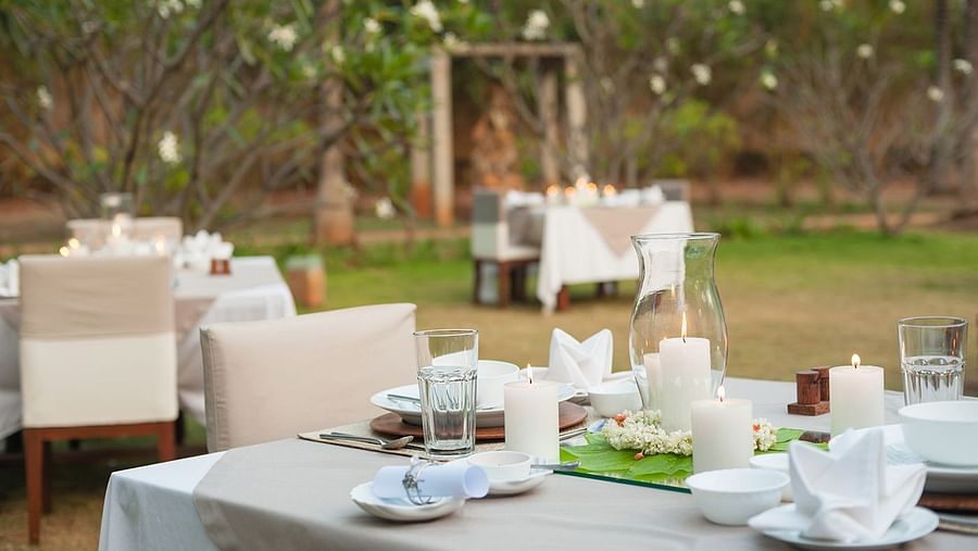 An outdoor dining setup with white tablecloths and chairs, green lawn and trees in the background - Shreyas Retreat