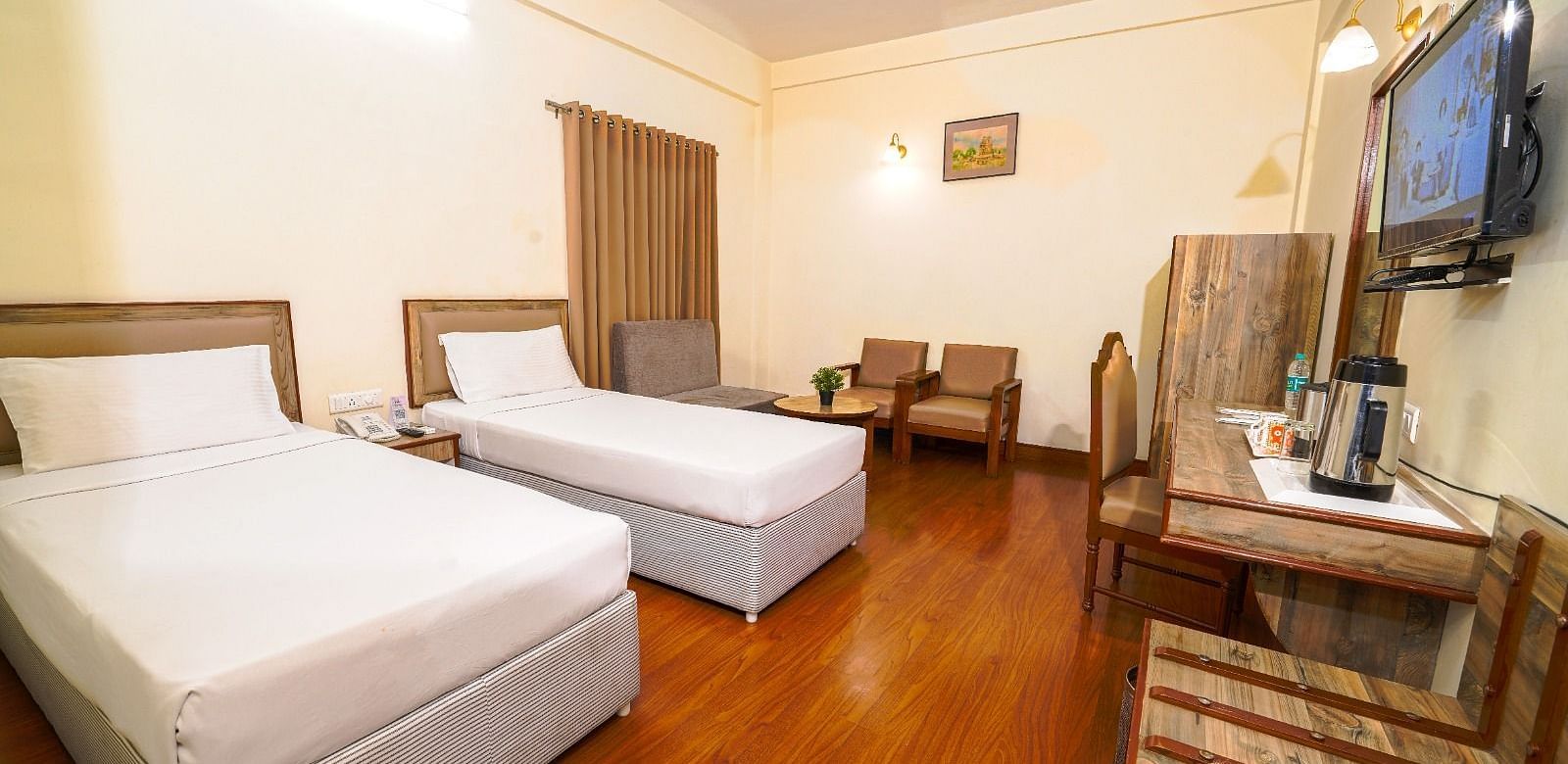 Well-lit hotel room with two single beds, wooden flooring, a spacious seating area, and a flat-screen TV in Hyderabad near Ramoji Film City.