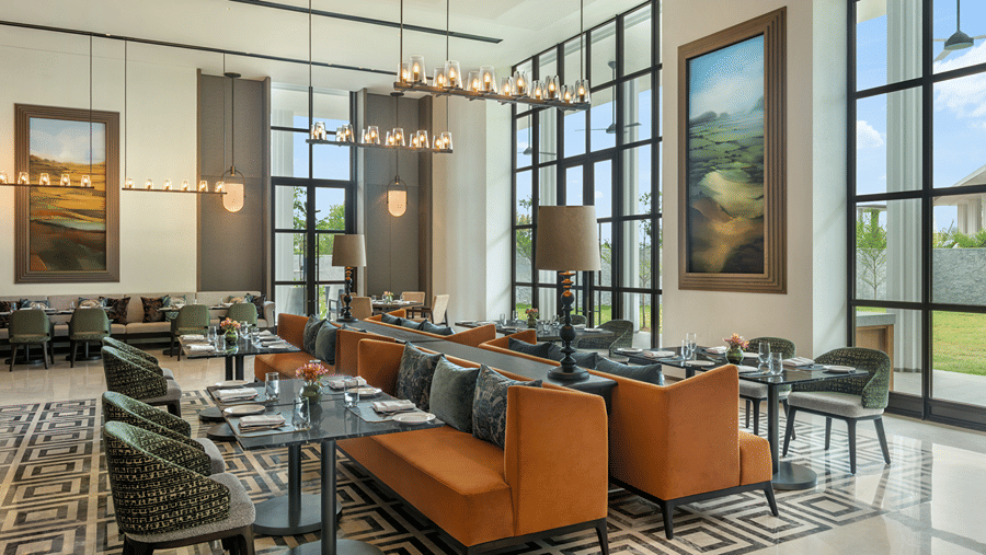Chithirai seating with orange banquettes, green patterned chairs, laid tables, landscape paintings and black-framed windows at The Residency Signature Madurai.