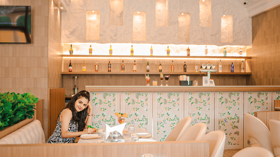 A woman sits at a table in the restaurant at Hotel Mumbai House Digha, Airoli, with a bar and hanging lights behind.