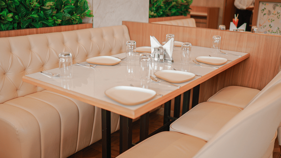 A set dining table with cream cushioned booths and green planters in the restaurant at Hotel Mumbai House Digha, Airoli.