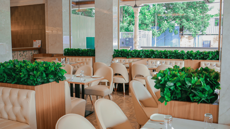 An indoor restaurant dining area with multiple tables, chairs, booths, and green planters at Hotel Mumbai House Digha, Airoli.