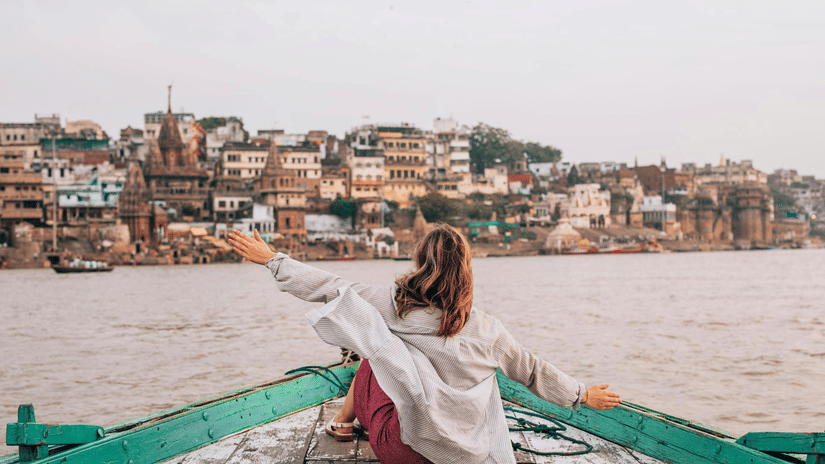A woman sitting on a wooden boat with arms outstretched, facing a riverfront city with buildings, temple structures, water in the foreground.