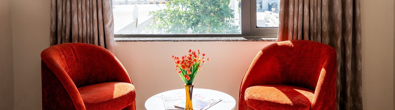 A cosy seating area at Daksh The Grand Gowardhan, Dwarka, with two vibrant red chairs by a window along with a coffee table, perfect for relaxation.
