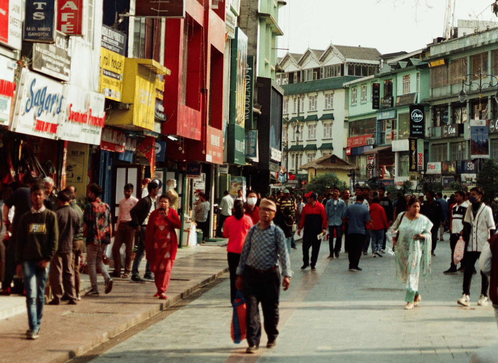 A crowd of people walking along a busy street lined with shops and multi-storey buildings on both sides.