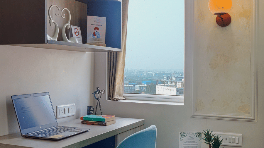 A study area at Urban Suites by BluSalzz, Jaipur, with a desk, a blue chair, bookshelves, and a window providing natural light.