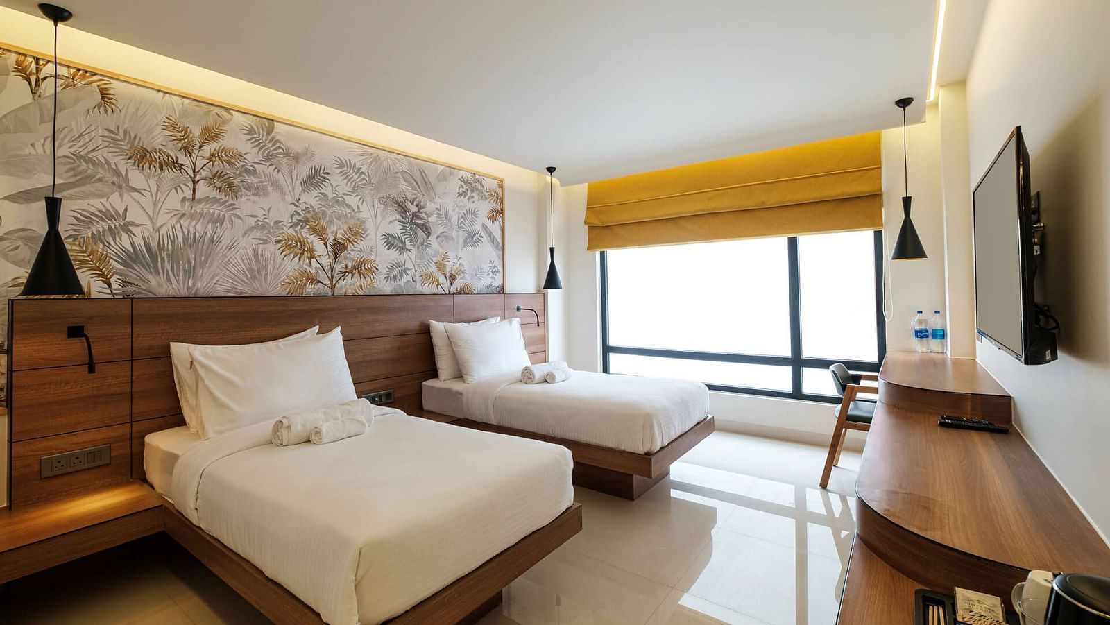 A spacious room at Indie Stays, Jaipur, with two beds and a joined wooden headboard, wall-mounted TV, wooden desk with a chair, and large alcove windows.